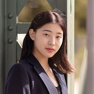 Portraitfoto von Dawon Yang. Sie trägt einen dunkelblauen Blazer über einem schwarzen Oberteil. Ihr schwarzes Haar fällt ihr über die rechte Schulter. Sie lehnt an einem grünen Stahlgerüst.