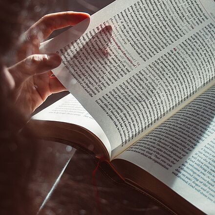 Ein aufgeschlagenes Buch wird in Händen gehalten. Das Lesezeichenband liegt auf der rechten Buchseite.