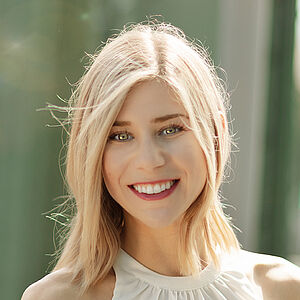 Portraitfoto von Melissa Panetta. Sie steht lächelnd auf einer grünen Stahlbrücke. Der Hintergrund ist verschwommen. Sie hat blondes schulterlanges Haar und trägt ein weißes Top. Sie lächelt mit geöffnetem Mund. 