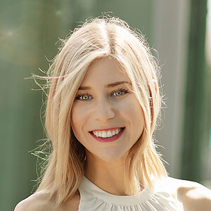Portraitfoto von Melissa Panetta. Sie steht lächelnd auf einer grünen Stahlbrücke. Der Hintergrund ist verschwommen. Sie hat blondes schulterlanges Haar und trägt ein weißes Top. Sie lächelt mit geöffnetem Mund. 