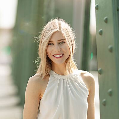 Portraitfoto von Melissa Panetta. Sie steht lächelnd auf einer grünen Stahlbrücke. Der Hintergrund ist verschwommen. Sie hat blondes schulterlanges Haar und trägt ein weißes Top. Sie lächelt mit geöffnetem Mund.