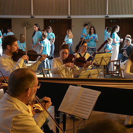 Im Vordergrund sitzt ein Orchester. Im Hintergrund steht eine große Gruppe Kinder (der Kinderchor). Alle Kinder tragen ein blaues Oberteil.