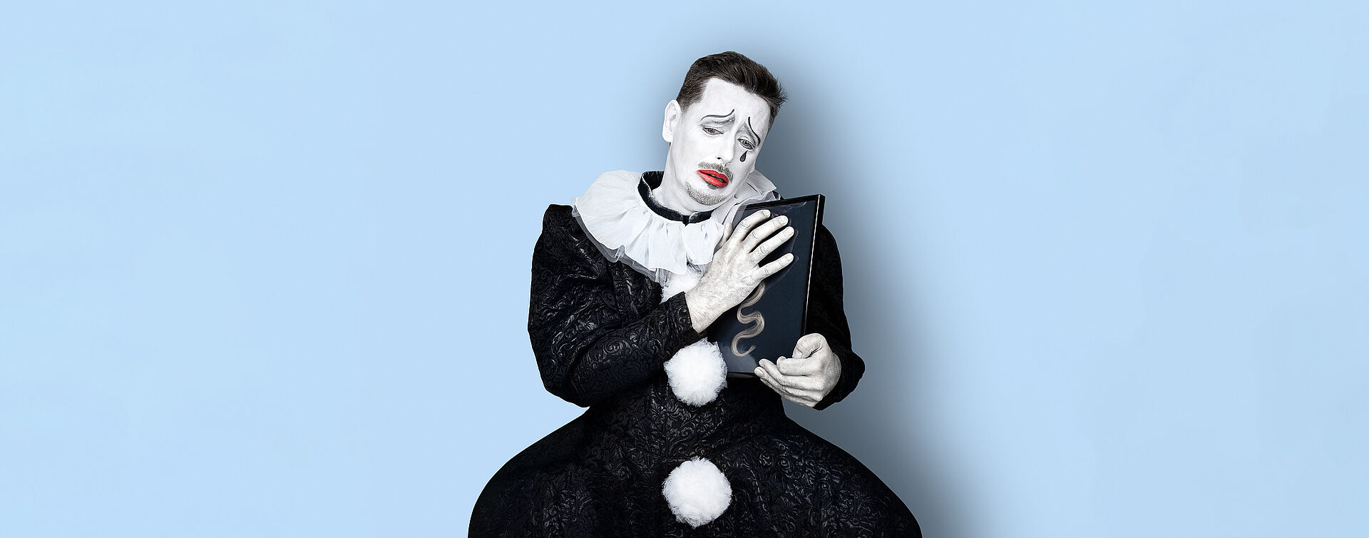 Ein Mann (Marcin Hutek) trägt ein schwarzes Clowns-Kostüm mit weißem Kragen und weißen Puscheln als Knopfleiste. Sein Gesicht ist weiß geschminkt, seine Lippen sind rot und eine schwarze Träne ist auf seiner Wange. In seinen Händen hält er einen Bilderrahmen mit einer blonden Locke, während er traurig schaut. Der Hintergrund des Bildes ist hellblau.