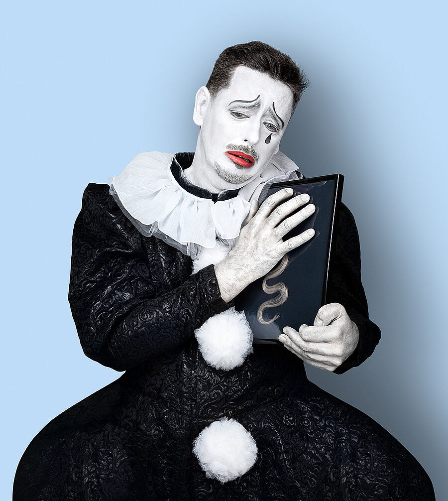 Ein Mann (Marcin Hutek) trägt ein schwarzes Clowns-Kostüm mit weißem Kragen und weißen Puscheln als Knopfleiste. Sein Gesicht ist weiß geschminkt, seine Lippen sind rot und eine schwarze Träne ist auf seiner Wange. In seinen Händen hält er einen Bilderrahmen mit einer blonden Locke, während er traurig schaut. Der Hintergrund des Bildes ist hellblau.