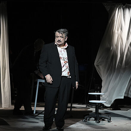 Im Vordergrund steht ein Mann. Im Hintergrund stehen Tische und Stühle. Zwei helle Vorhänge hängen bis zum Boden.