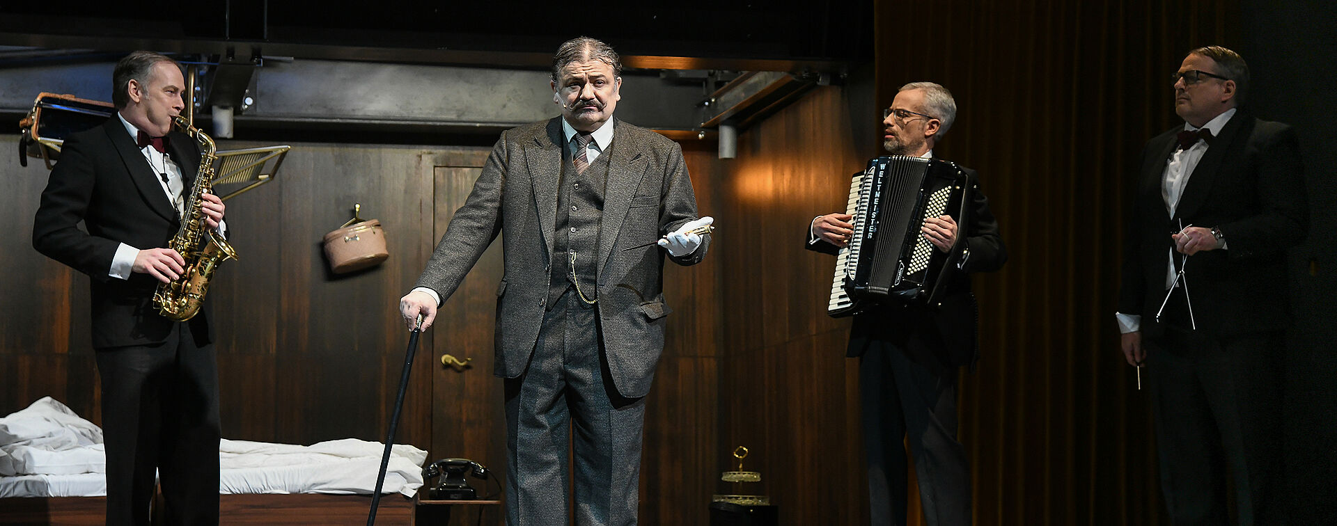 Hercule Poirot steht mit einem Messer in der Hand da, über den ein weißer Handschuh gestülpt ist. Um ihn herum ein Saxophonist, ein Akkordeonist und ein Triangelspieler.