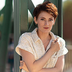 Portraitfoto von Lidia Melnikova. Sie lehnt an grünen Stahlstreben. Ihren linken Arm hält sie unter der Brust, der rechte Ellbogen stützt sich darauf auf. Ihr rötlich-braunes Haar ist kurz und zum Seitenscheitel gestylt. Sie trägt eine beige-karierte Bluse, eine dezente Kette, kleine Creolen und einen silbernen Ring.